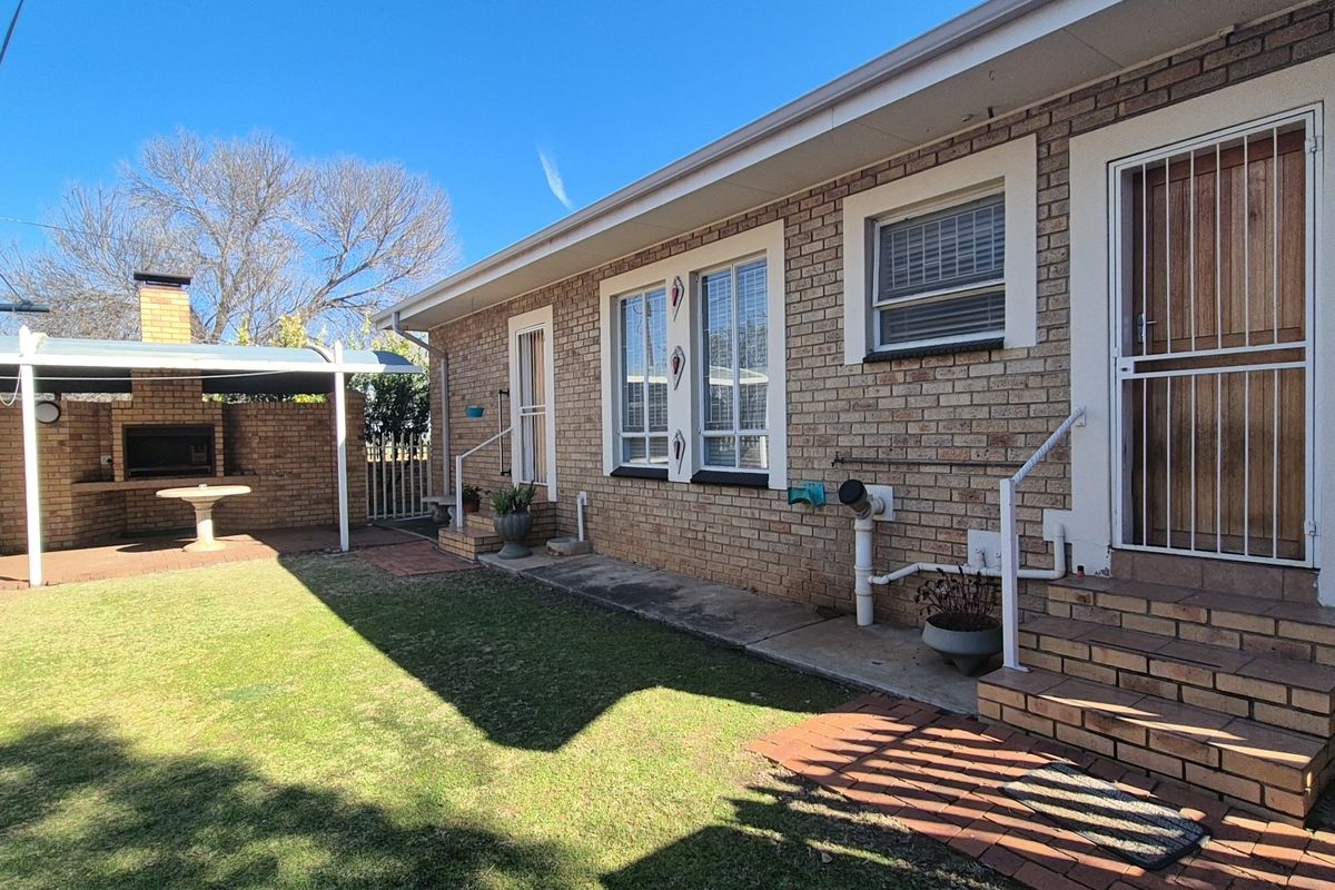 Rear exterior view featuring a brick facade, paved walkway, and a covered outdoor braai area set within a garden space.
