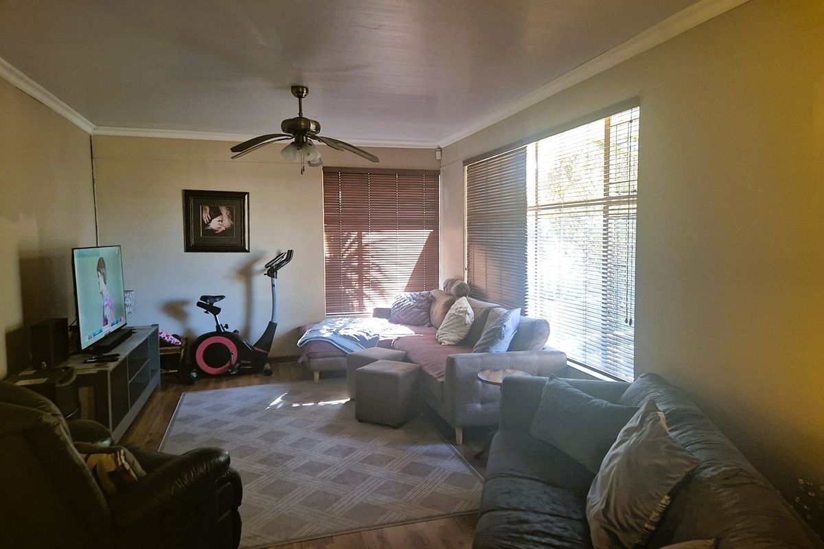 A comfortable living room featuring laminate flooring, a ceiling fan, and large windows with venetian blinds, providing a functional and well-lit space.