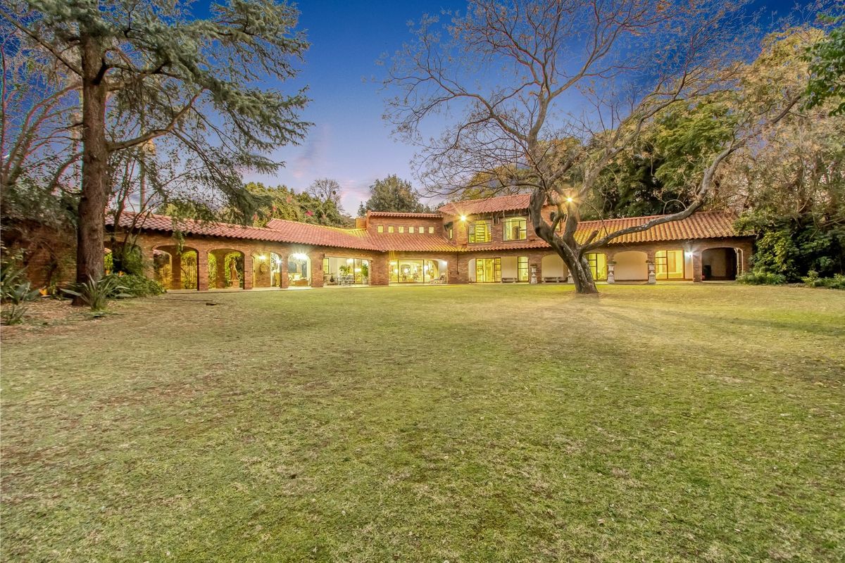 Spacious rear garden view featuring a large lawn, mature trees, and a distinctive brick residence with a terracotta tile roof and arched colonnade.