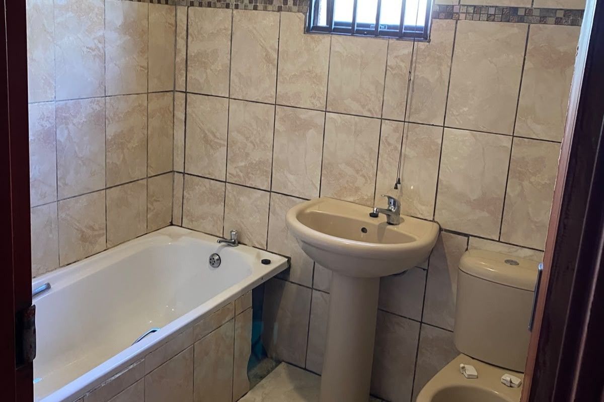 A functional bathroom featuring a bathtub, pedestal basin, and toilet with full-height wall tiling and a small window with security bars.