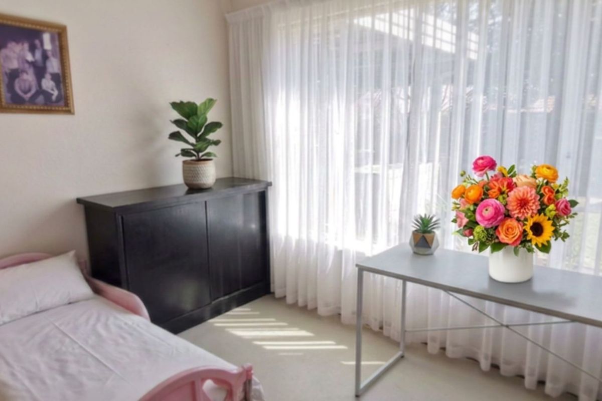 A bright bedroom featuring neutral walls, carpet flooring, and large windows dressed with sheer curtains, providing ample natural light.