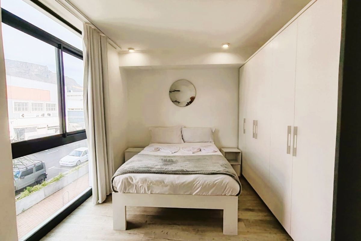 A bright and modern bedroom featuring built-in floor-to-ceiling cupboards, laminate flooring, and a large window offering natural light and urban views.