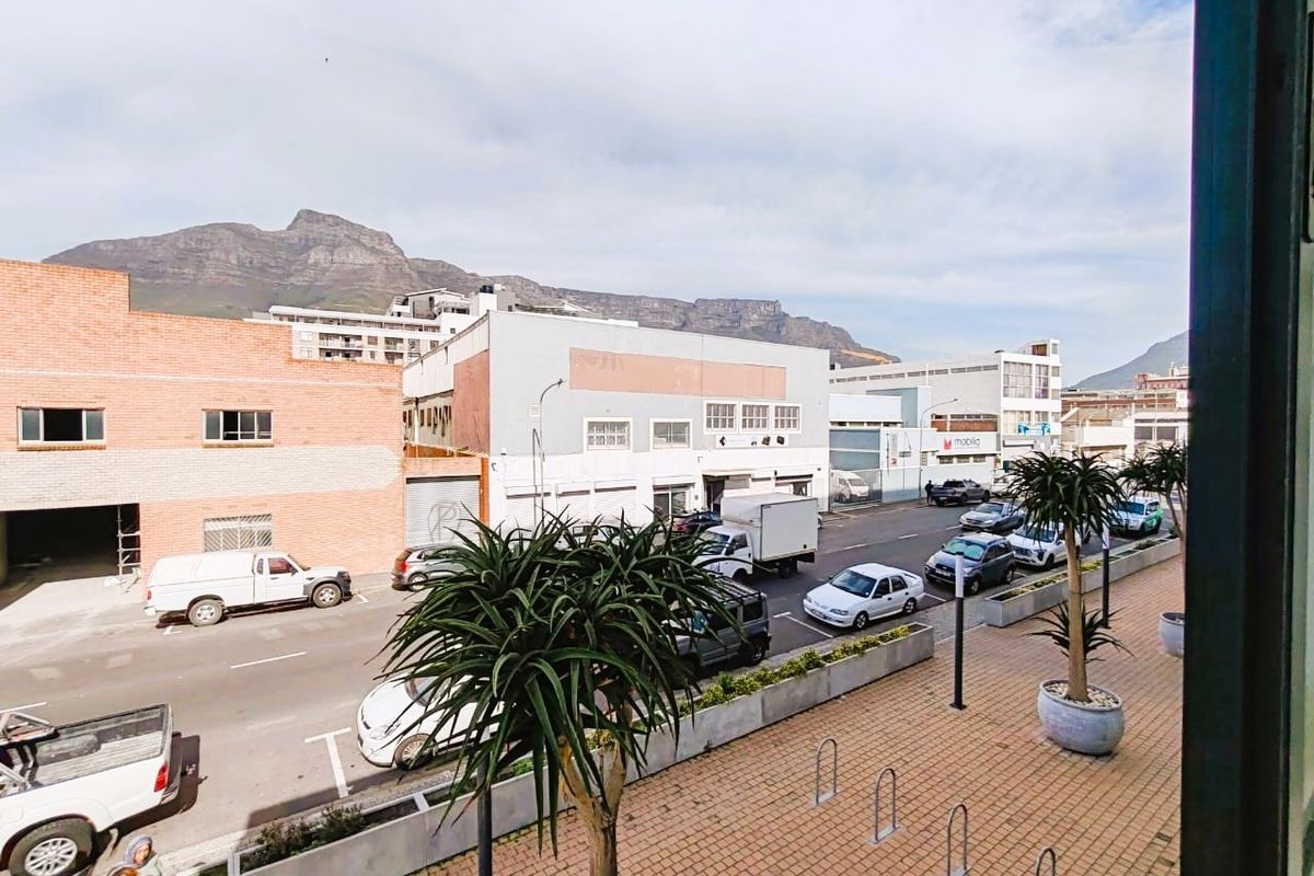 An elevated street-level view overlooking a paved sidewalk with bicycle racks, planters, and surrounding urban buildings with mountain views in the distance.