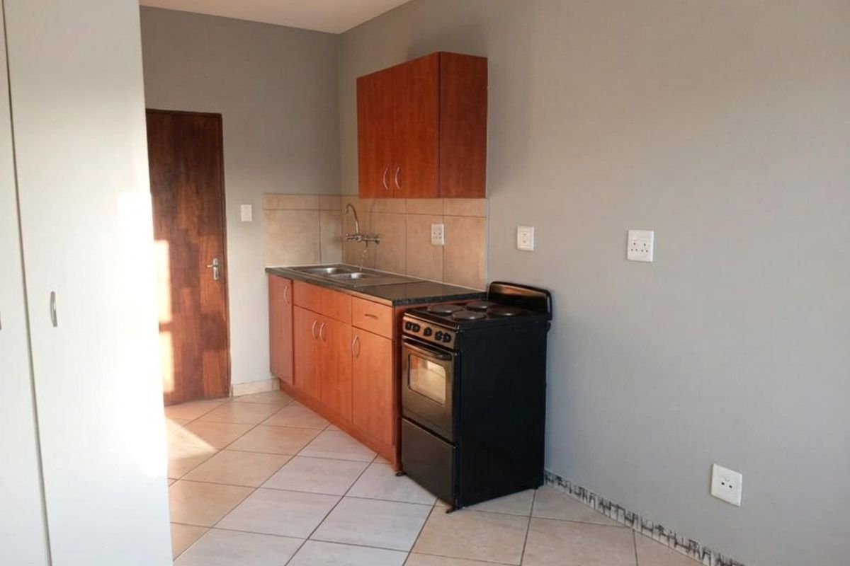 A compact kitchen featuring wooden cabinetry, a stainless steel sink, tiled backsplash, and a freestanding electric stove with tiled flooring.