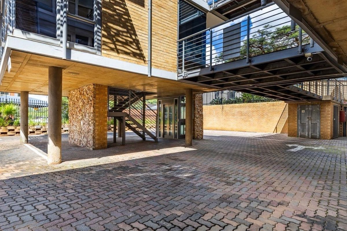 A modern commercial exterior courtyard featuring brick paving, stone-clad structural columns, an elevated steel walkway, and a glass elevator access point.
