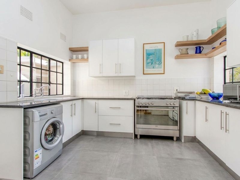 Modern kitchen featuring white cabinetry, granite countertops, stainless steel gas stove, tiled backsplash, and floating timber shelving with large steel-framed windows.