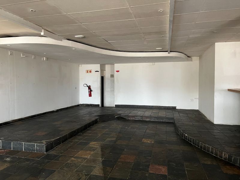 An empty open-plan restaurant space featuring a raised platform area, dark tiled flooring, and a suspended ceiling with recessed lighting.