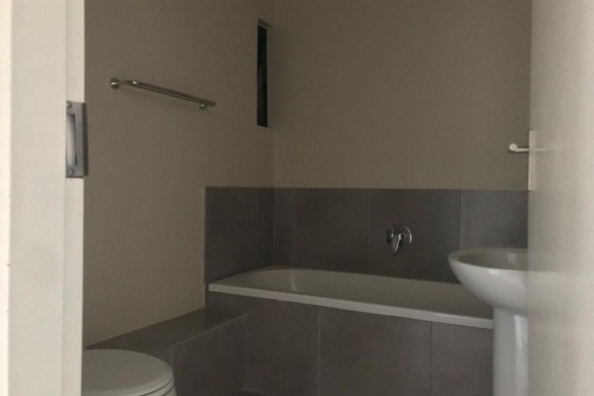 A compact bathroom featuring a built-in bathtub, pedestal basin, and toilet with neutral grey wall tiling.