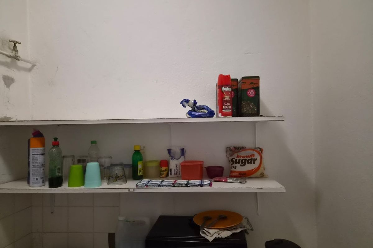 A basic kitchen storage area featuring wall-mounted shelving, a water tap, and tiled splashback, showing signs of wear and dated finishes.
