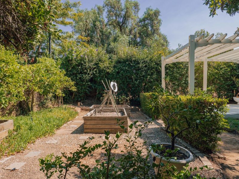 A well-maintained garden featuring a timber pergola, gravel pathways, and raised timber garden beds surrounded by established hedging and mature trees.