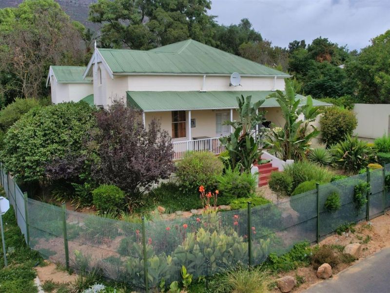 A charming single-story house featuring a green corrugated metal roof, a covered front veranda with timber railings, and a well-established garden.