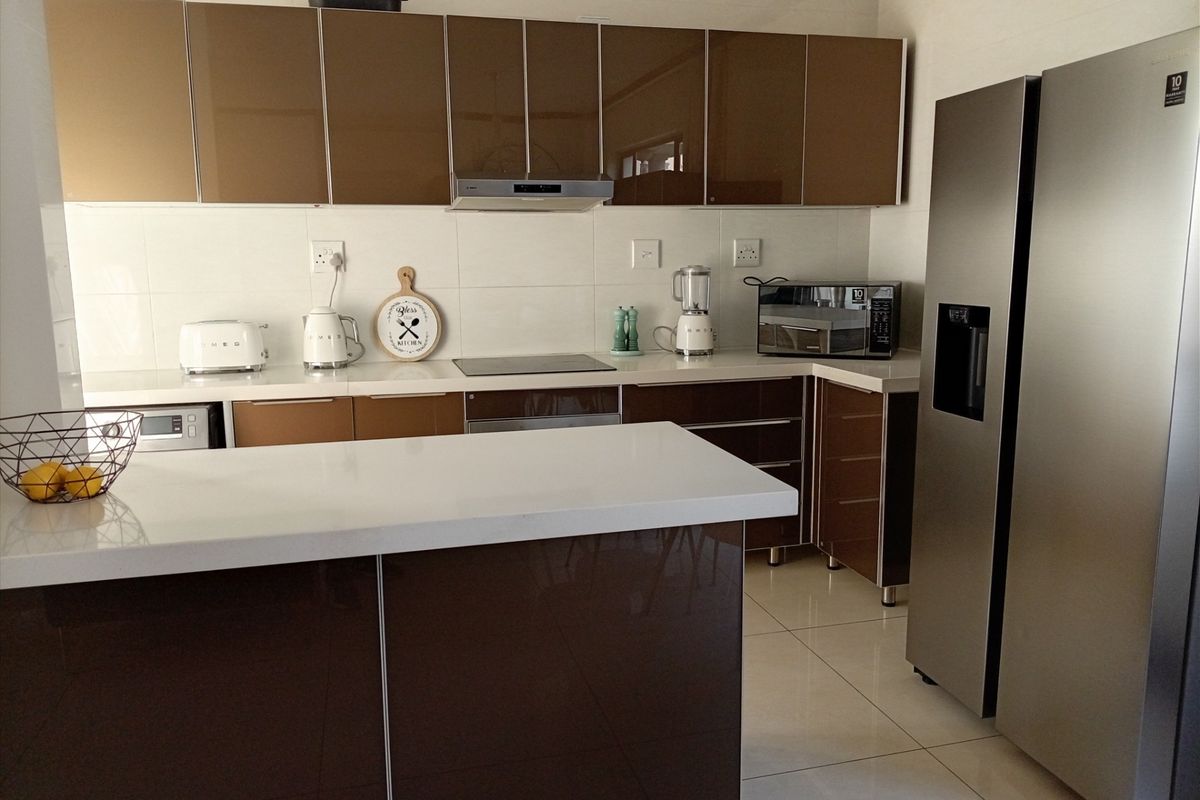 Modern kitchen featuring high-gloss brown cabinetry, white stone countertops, a central island, and integrated appliances with tiled flooring.