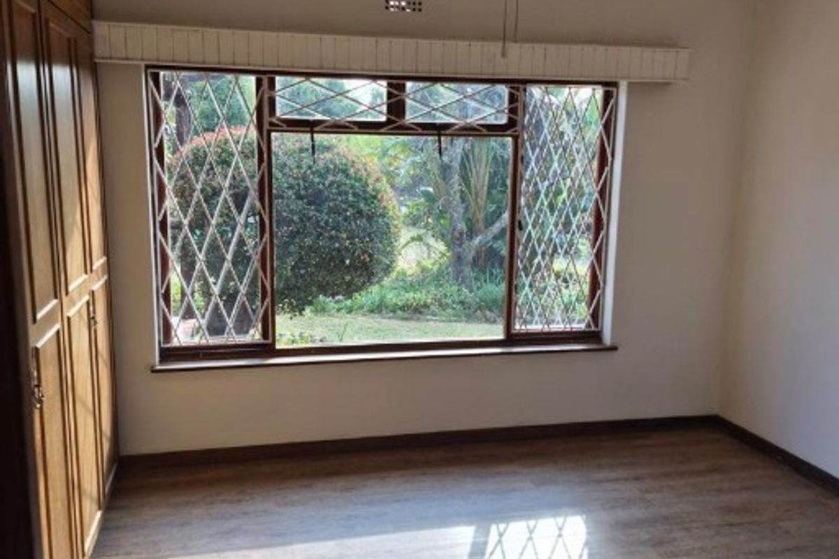 A vacant bedroom featuring timber-look flooring, built-in cupboards, a ceiling fan, and large windows with security bars overlooking the garden.
