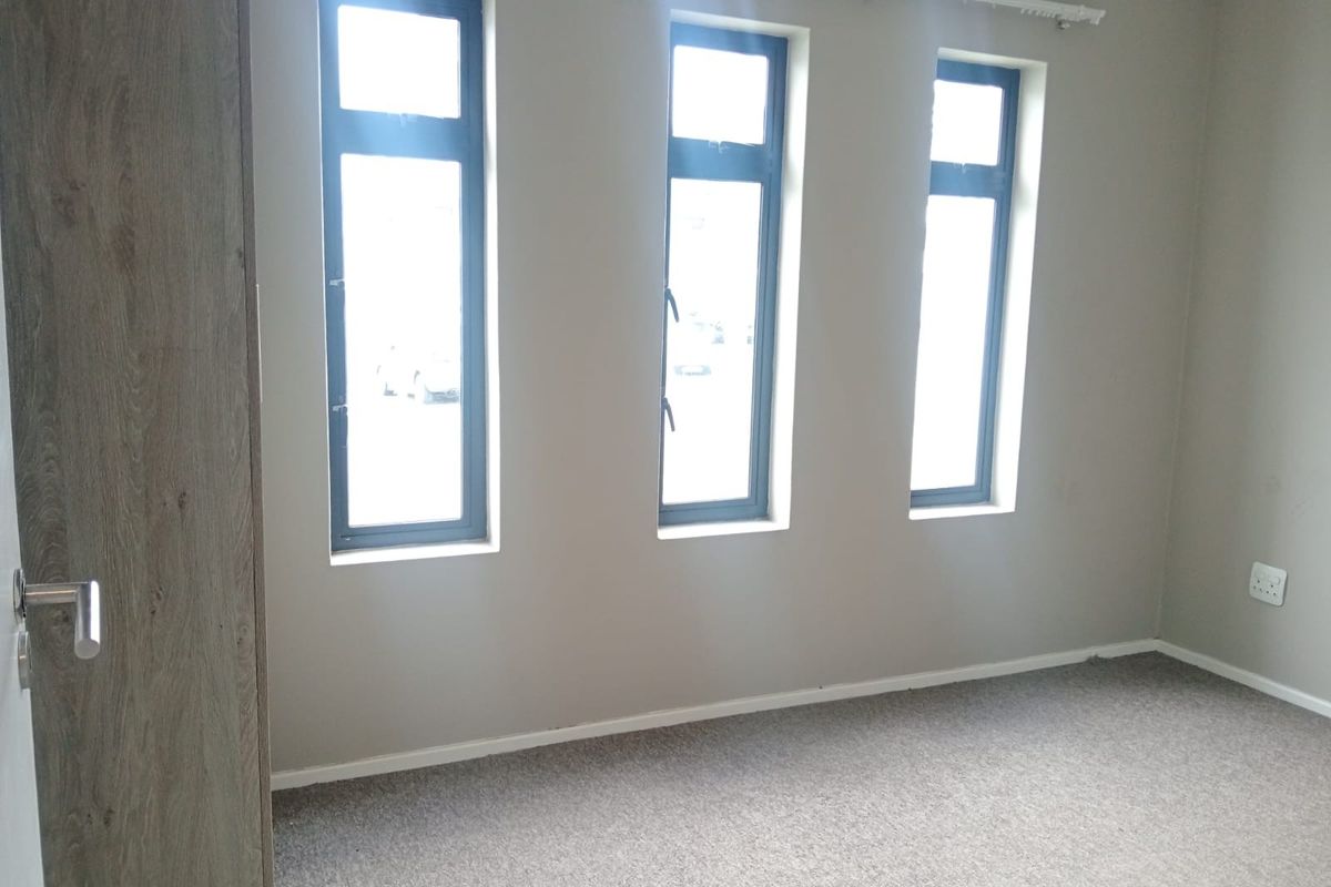 A bright, empty bedroom featuring neutral walls, carpeted flooring, built-in cupboard storage, and three tall windows with modern aluminium frames.