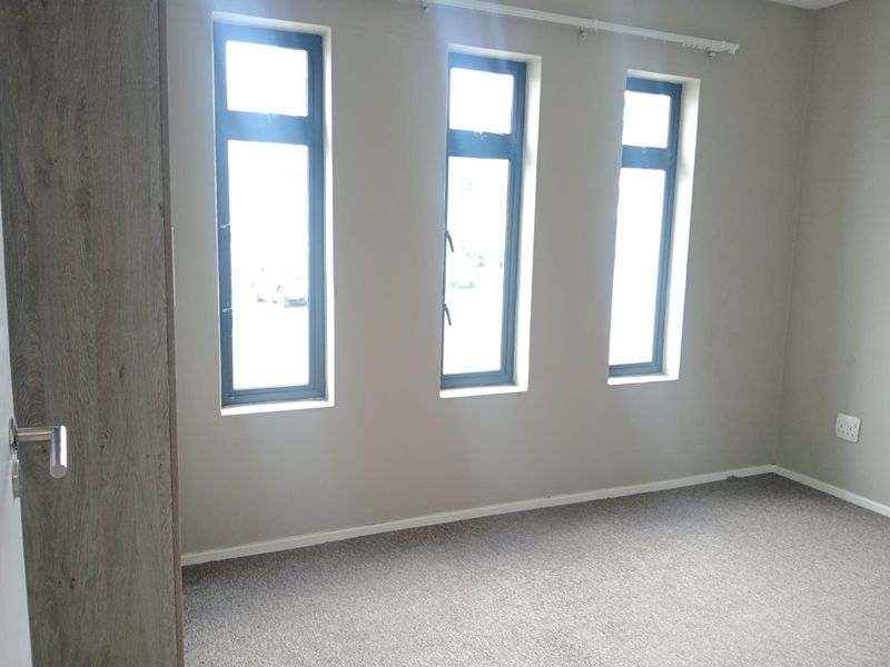 A bright, empty bedroom featuring neutral walls, carpeted flooring, built-in cupboard storage, and three tall windows with modern aluminium frames.