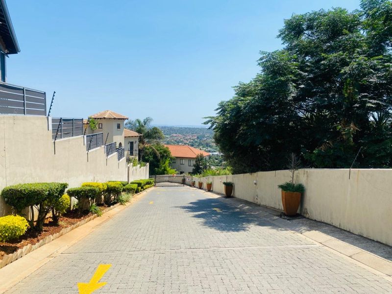 A paved driveway leading to a secure gated entrance, featuring perimeter walls with electric fencing and views of the surrounding neighborhood.