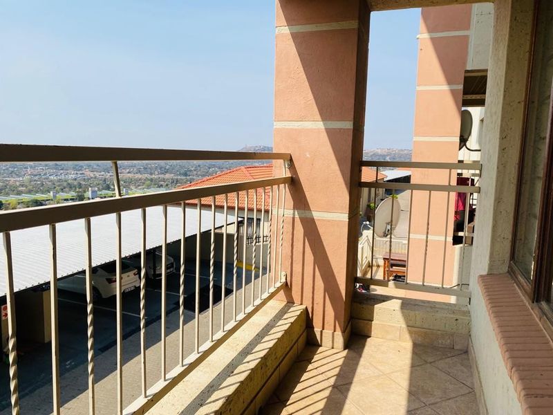 A covered balcony featuring tiled flooring and a metal balustrade, offering elevated views over the complex parking area and surrounding landscape.