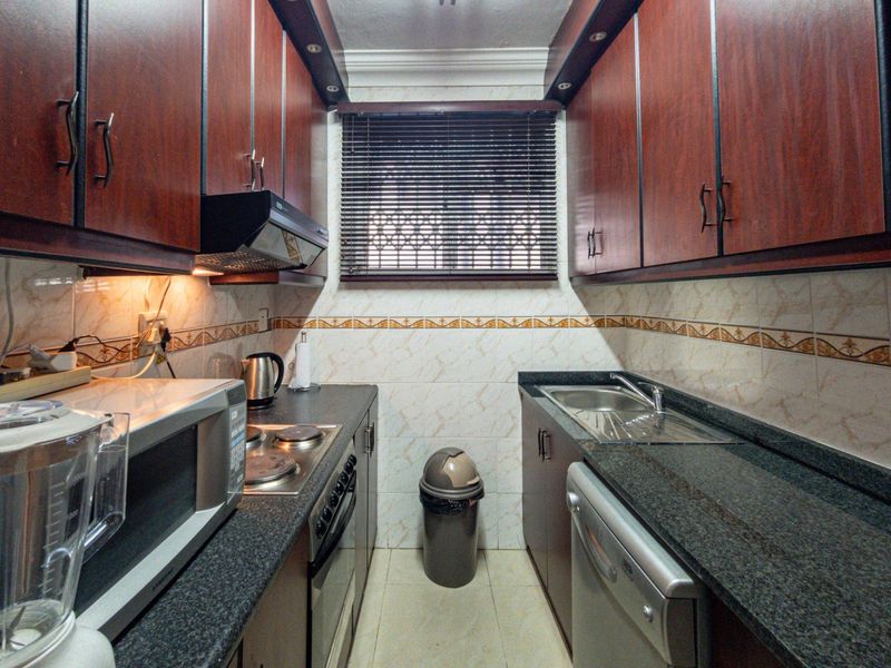 A compact galley-style kitchen featuring dark wood-look cabinetry, granite countertops, integrated oven and hob, and tiled wall finishes.