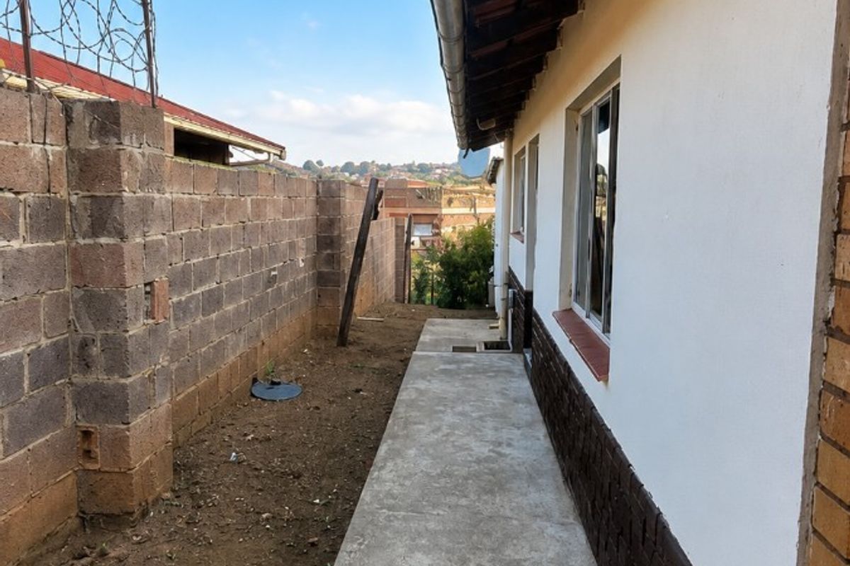 A side exterior view of the property featuring a concrete walkway, brick boundary wall with security fencing, and exposed roof eaves.