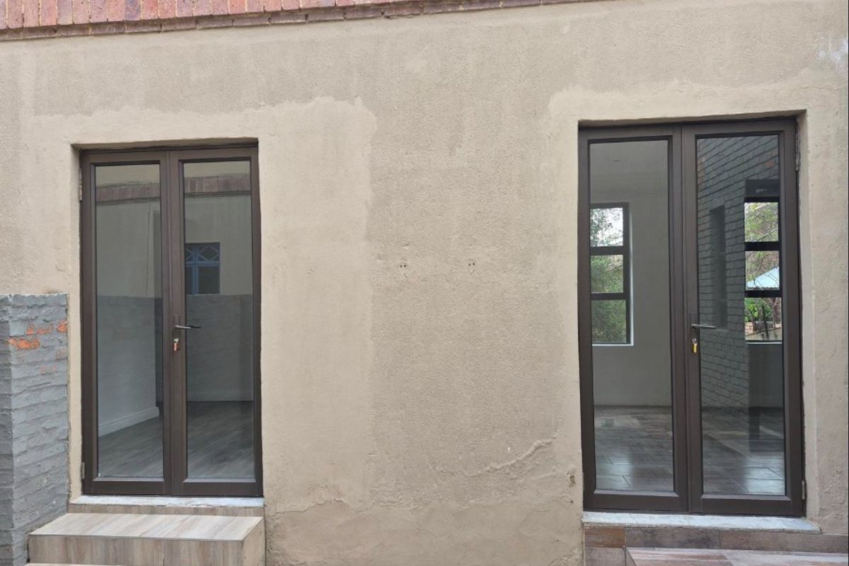 Rear exterior view featuring two sets of modern aluminium framed glass double doors with tiled steps and unfinished plastered wall surfaces.