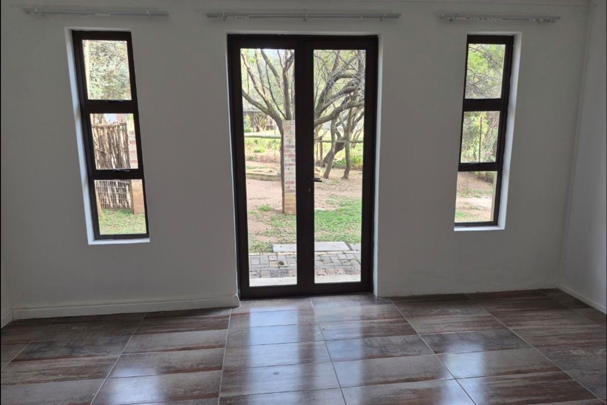 A bright, empty living room featuring modern wood-look tiled flooring, double glass doors, and side windows providing a view of the garden.