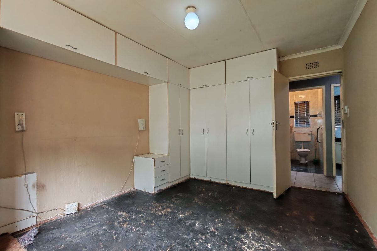 An empty bedroom featuring built-in cupboards and overhead storage, with direct access to an en-suite bathroom. The room requires flooring and wall finish updates.