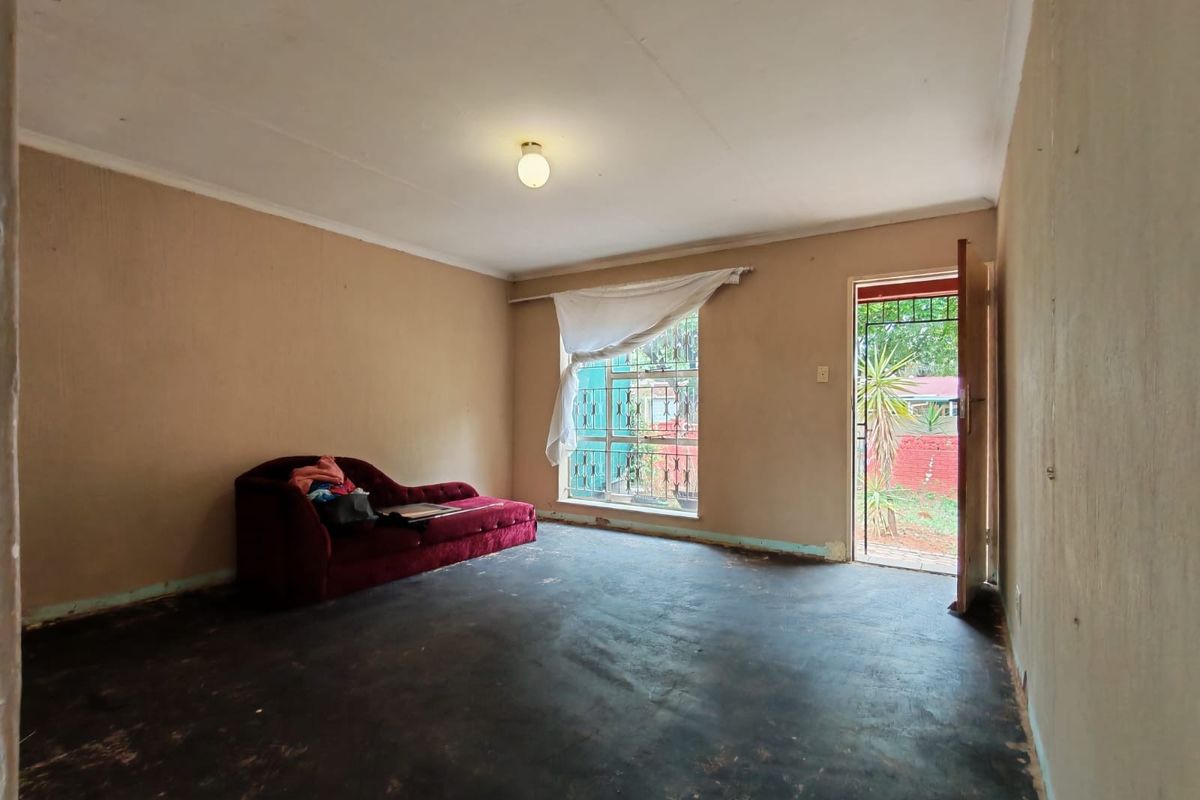 A living room space featuring a security gate, metal window frame, and ceiling cornice, currently requiring significant cosmetic updates and flooring replacement.
