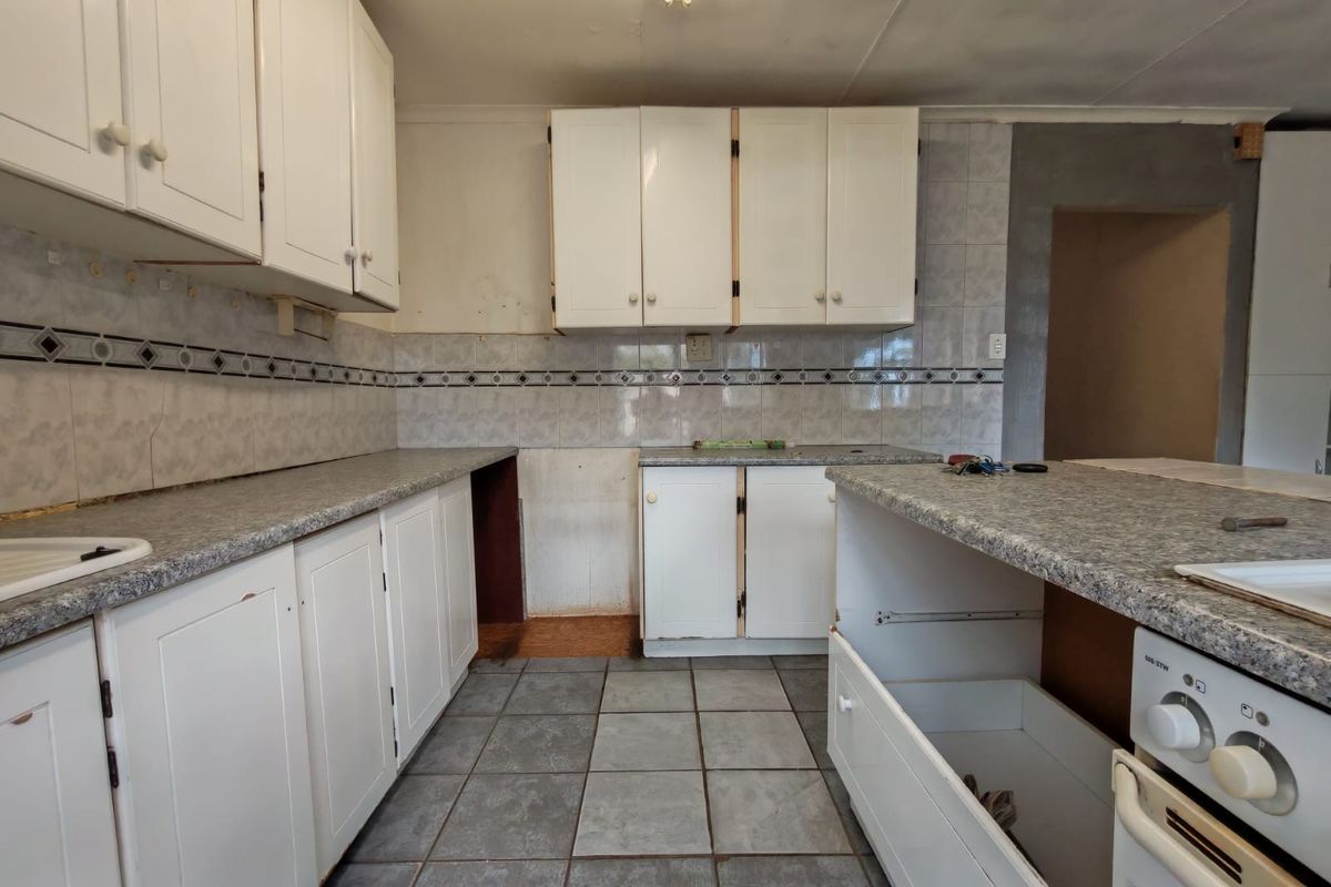 A dated kitchen featuring white cabinetry, laminate countertops, and tiled flooring, currently requiring renovation and completion of missing fixtures.