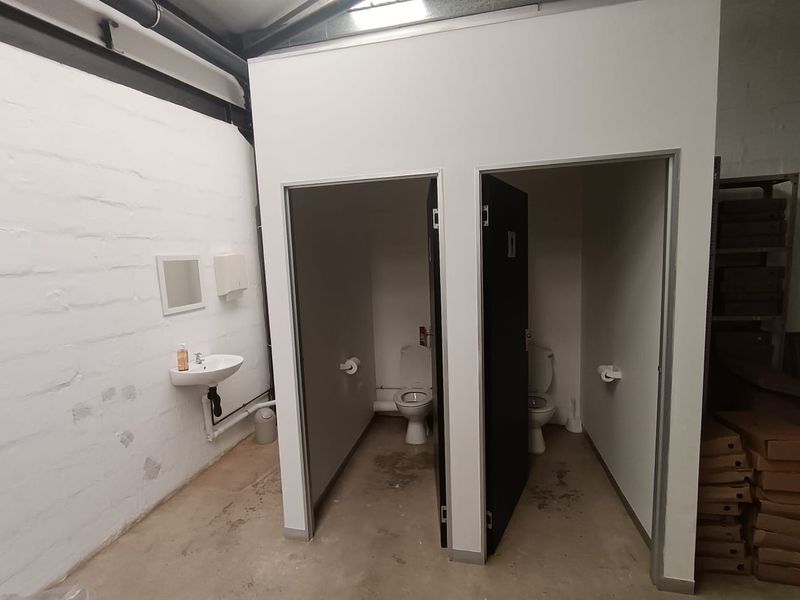 Commercial bathroom facility featuring two private toilet cubicles and a wall-mounted basin within an industrial-style space with exposed ceiling and painted brick walls.