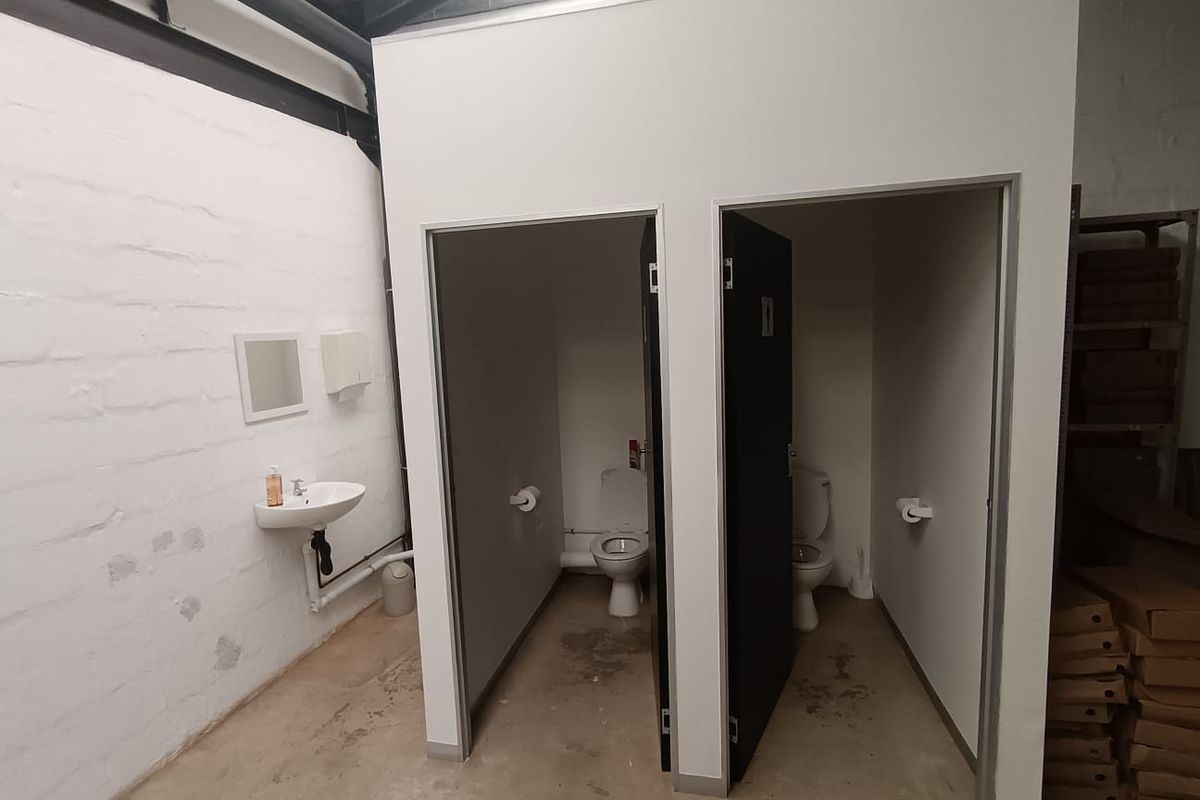 Commercial bathroom facility featuring two private toilet cubicles and a wall-mounted basin within an industrial-style space with exposed ceiling and painted brick walls.