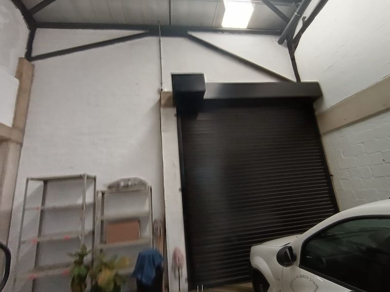 Commercial garage space featuring high ceilings with exposed steel trusses, a large roller shutter door, and natural light from a skylight.