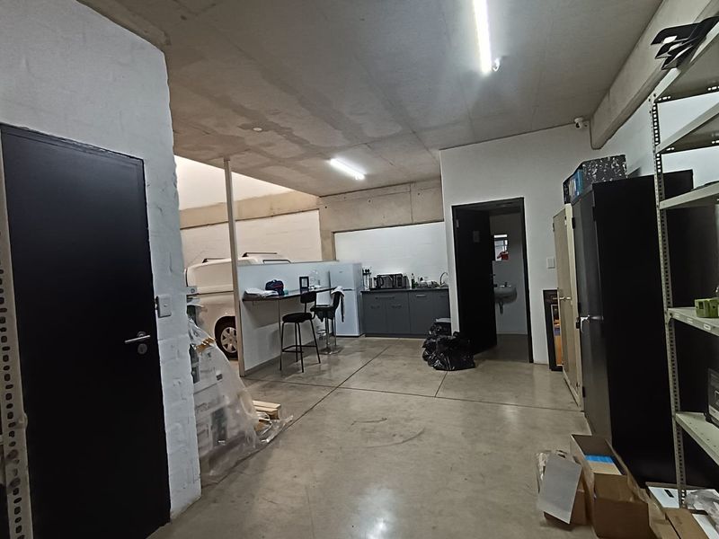 A functional commercial utility space featuring concrete flooring, exposed ceiling, and a small kitchenette area with sink and cabinetry.