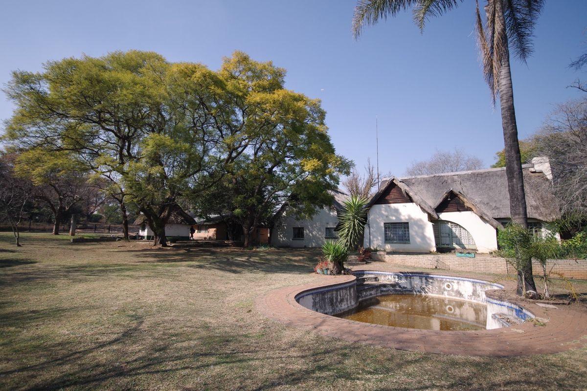Exterior view of a farm property featuring a thatched-roof dwelling and a swimming pool requiring maintenance, set within a large garden area.