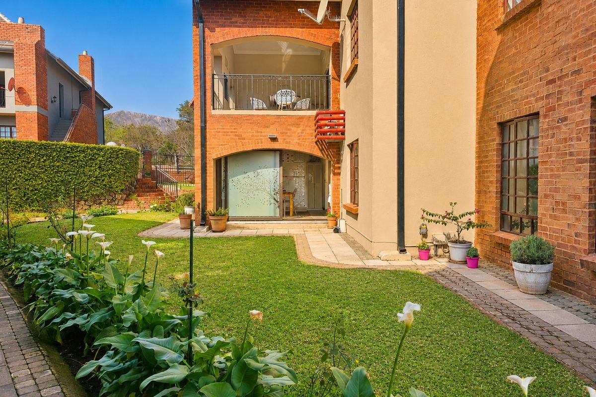 Rear view of the apartment building featuring a private garden, paved walkway, and a covered balcony with metal railings.