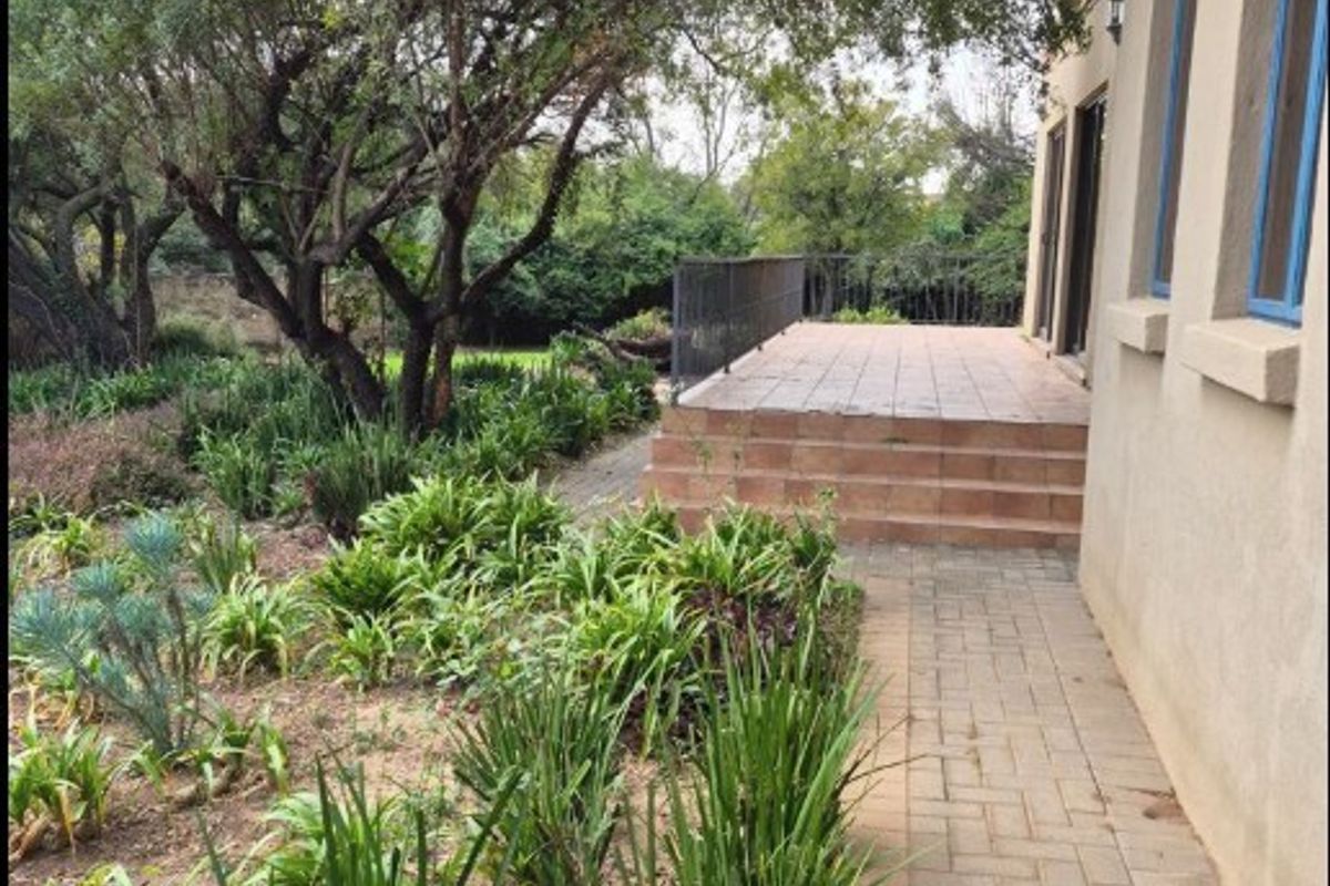A rear exterior view featuring a tiled patio with metal railing, a paved walkway, and a mature landscaped garden.