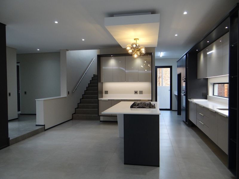 Modern, spacious kitchen featuring a central island with gas cooktop, sleek built-in cabinetry, integrated appliances, and recessed lighting, adjacent to an internal staircase.