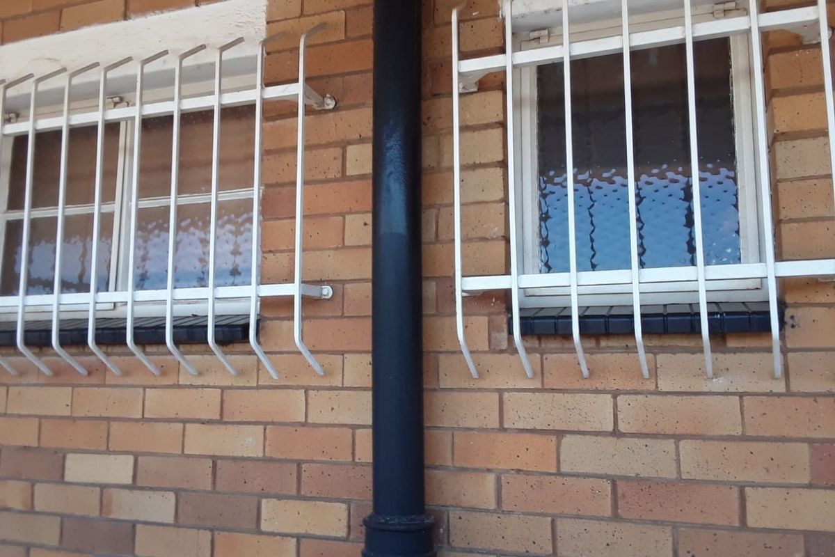 Exterior view of a brick apartment facade featuring two windows with white security bars and a central downpipe.