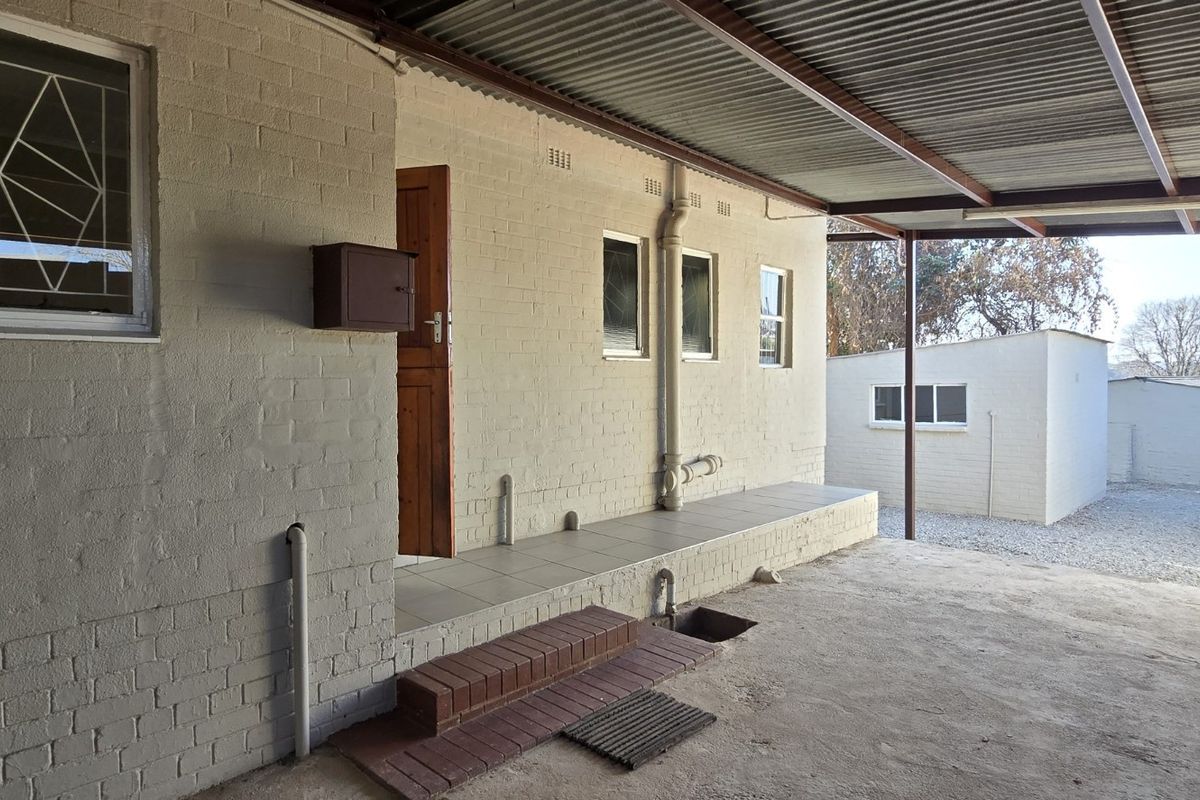 A covered rear patio area featuring a brick facade, tiled entrance step, and corrugated metal roofing, providing access to the rear yard and outbuilding.