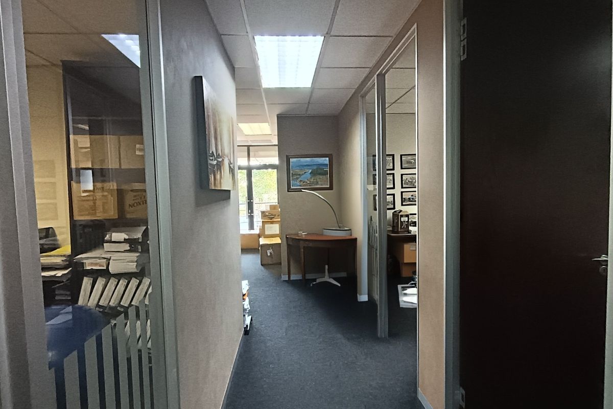 A functional office hallway featuring glass partitions, suspended ceilings with recessed lighting, and carpeted flooring, providing access to adjacent office spaces.