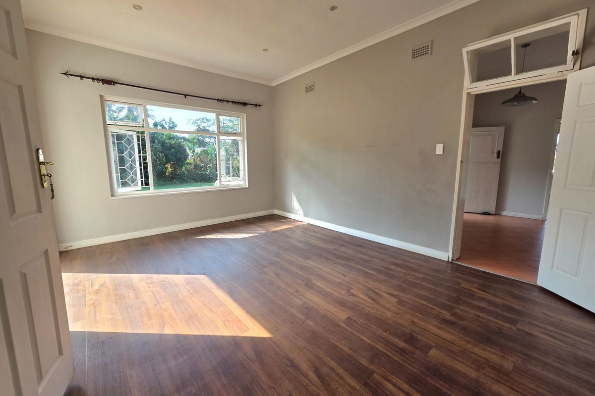 A bright and spacious living room featuring warm-toned laminate flooring, recessed lighting, and a large window overlooking the garden.