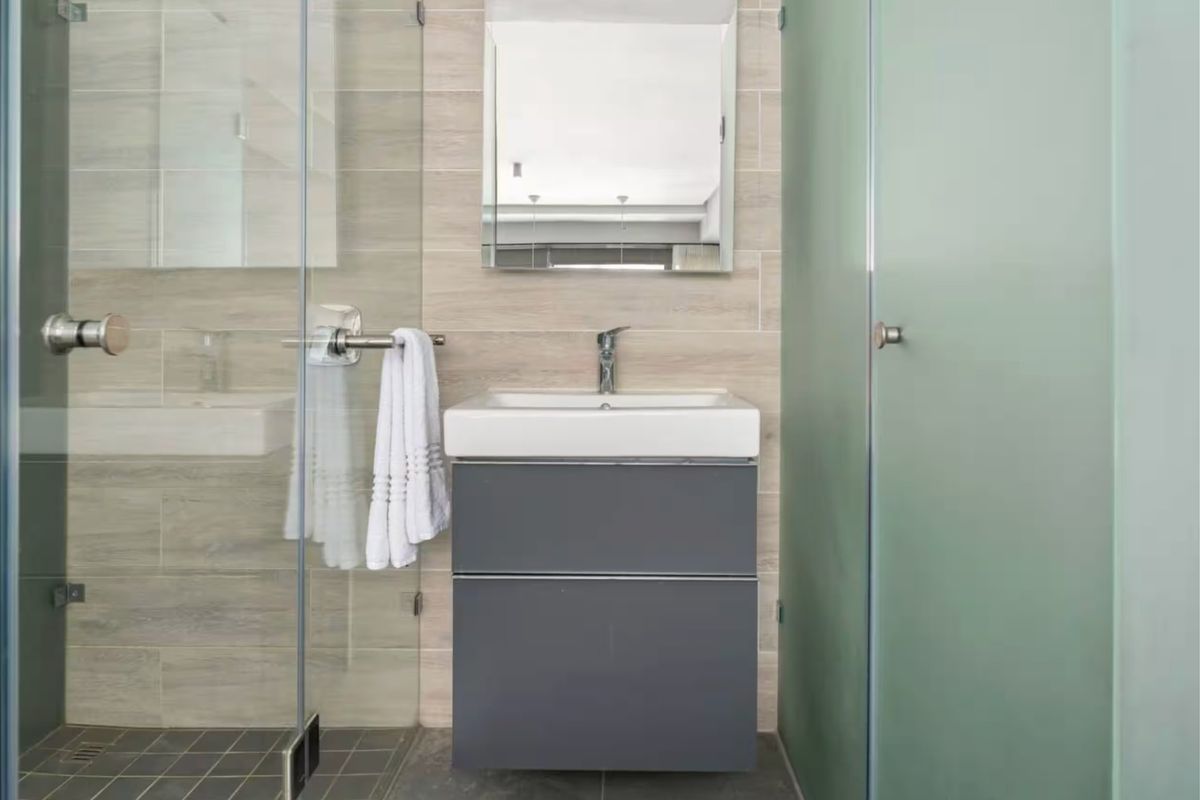 Modern bathroom featuring a sleek grey vanity cabinet, ceramic basin, wall-mounted mirror, and a glass-enclosed shower with neutral tiled finishes.