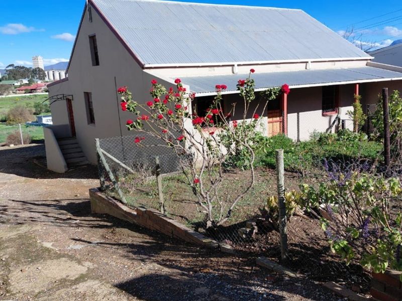 A detached house featuring a corrugated metal roof and a covered front veranda, set on a plot with a garden and chain link fencing.