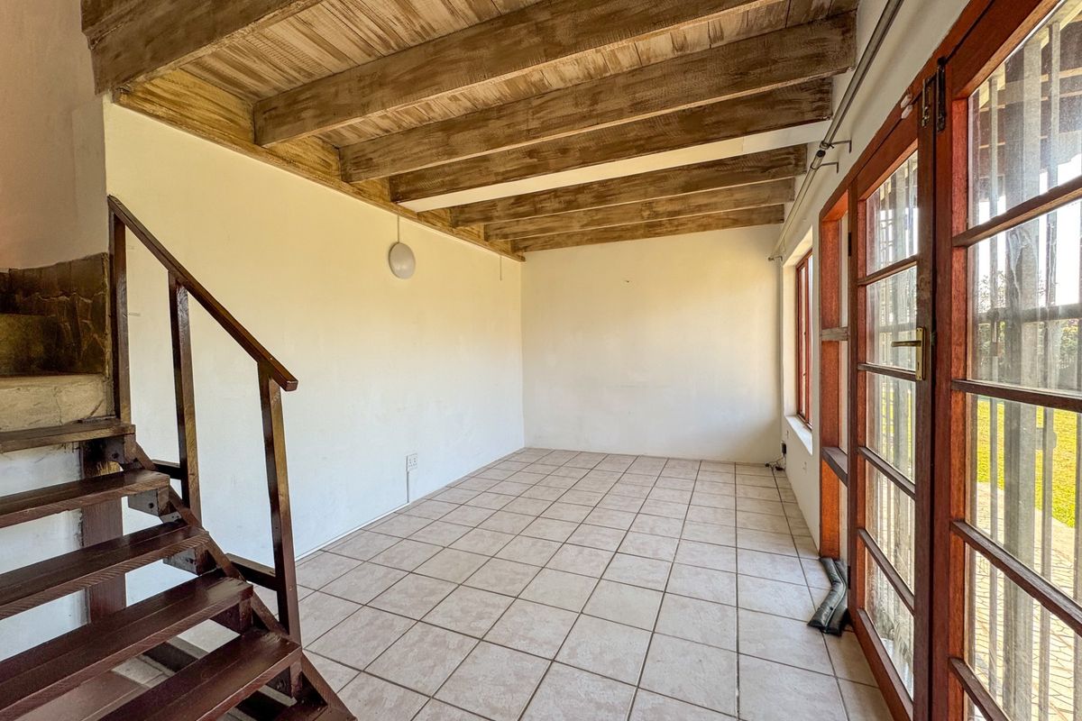 An empty living area featuring exposed timber ceiling beams, ceramic floor tiles, and a timber staircase with direct access to the garden.