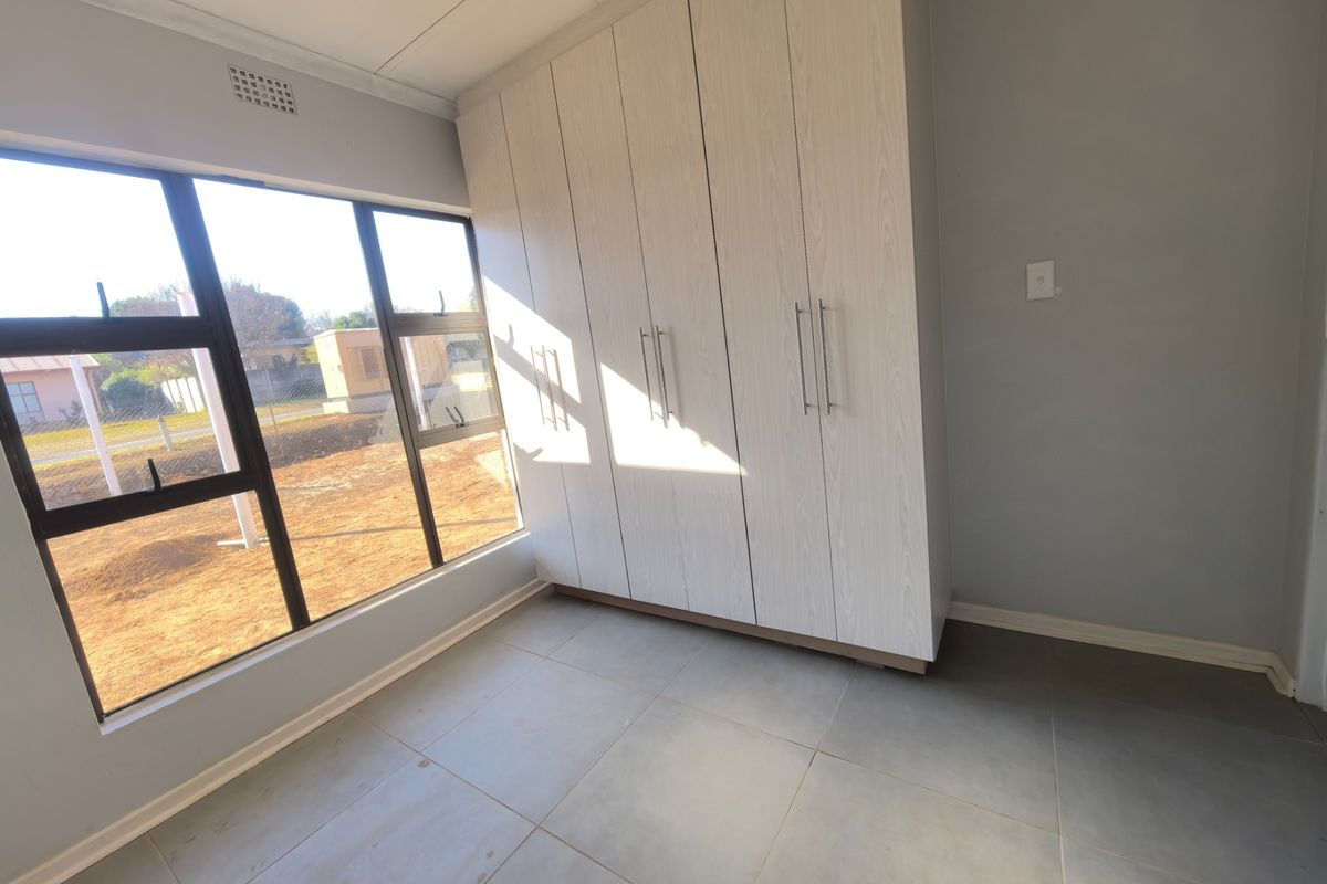 A bright, empty bedroom featuring modern light-wood built-in cupboards, large aluminium-framed windows, and neutral grey floor tiles.