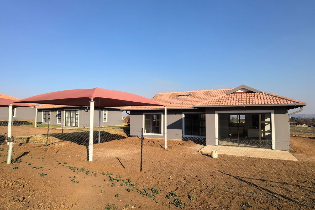 Rear view of a modern house featuring a tiled roof with solar geyser, a concrete patio, and a shade cloth carport in an unlandscaped yard.