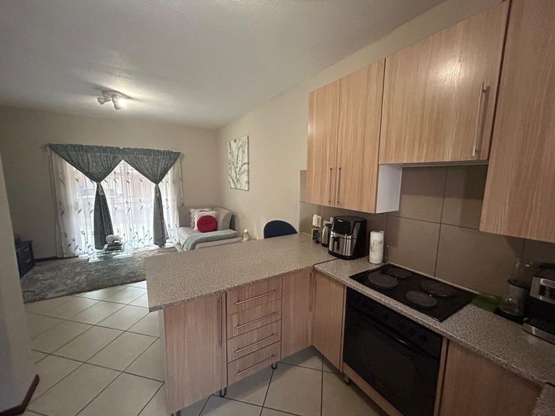 Open-plan living area featuring a functional kitchen with light wood cabinetry, electric hob, and tiled flooring, flowing into a bright lounge with sliding door access.
