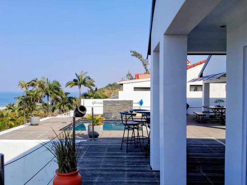 Spacious covered patio featuring timber decking and glass balustrades, overlooking a private swimming pool with scenic ocean views.