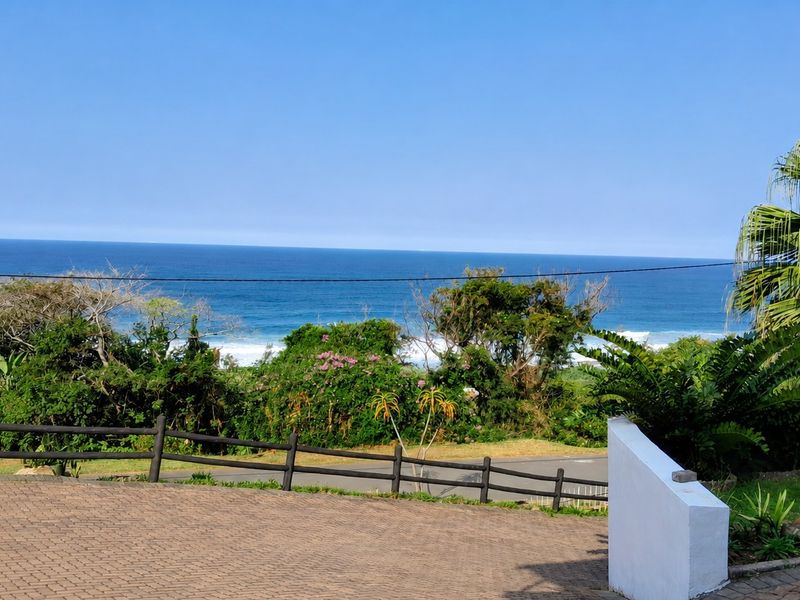 A scenic elevated view overlooking the ocean, featuring a paved driveway, landscaped garden area, and timber boundary fencing.