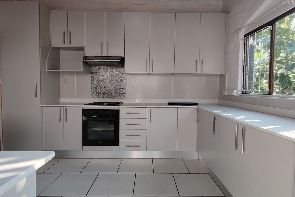 A clean, modern kitchen featuring white cabinetry, built-in oven and hob, tiled splashback, and large windows with security bars.