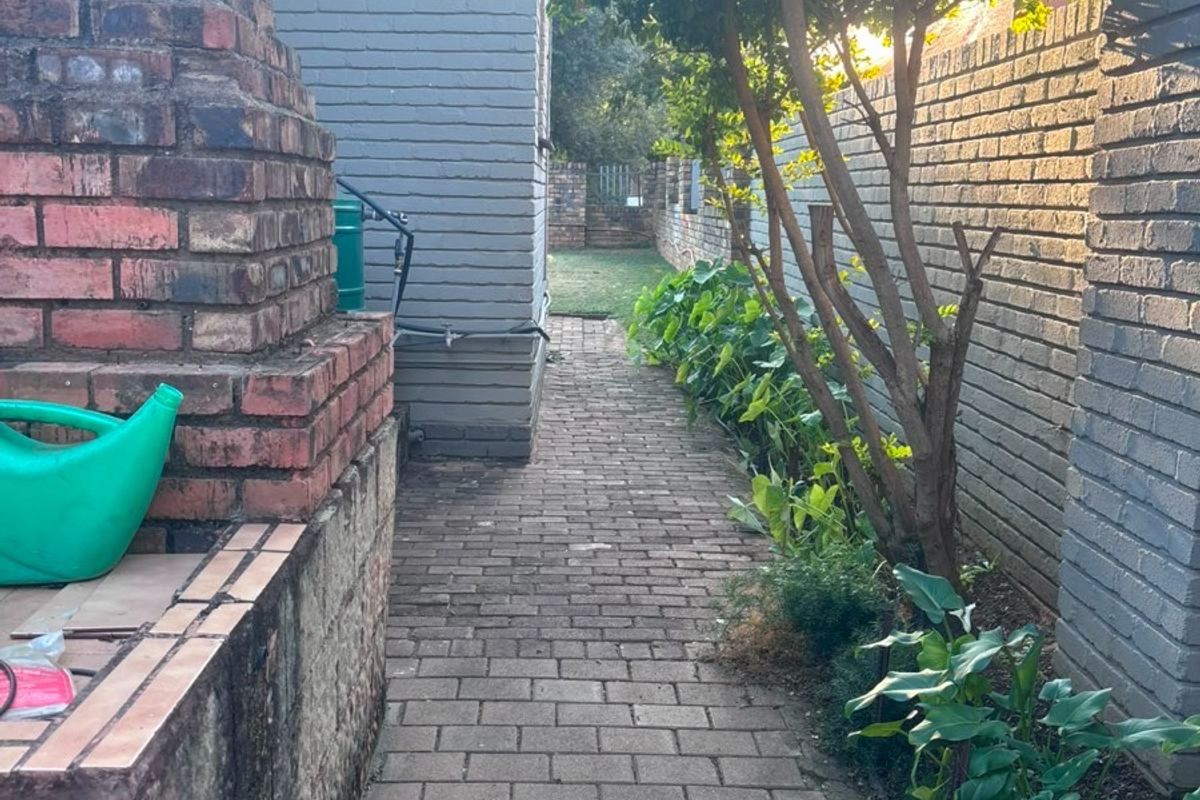 A narrow rear garden pathway featuring brick paving, a built-in brick fireplace, and perimeter boundary walls.